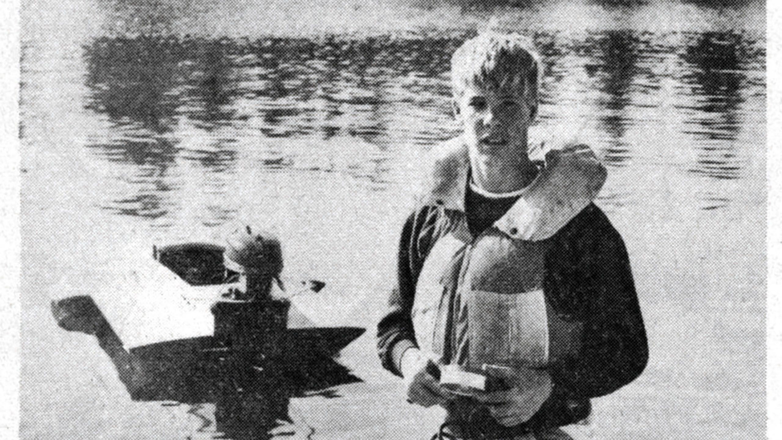 Black and white photo of a young Miniea wearing a lifeboat jacket with a motorboat behind him